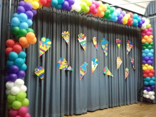 Bunte Luftballons und festliche Banner schmücken einen Vorhang in einem Veranstaltungsraum.